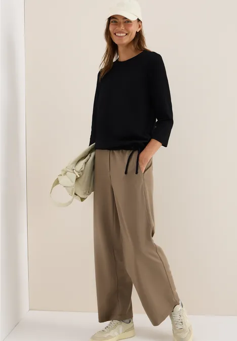 Frau mit schwarzem Oberteil, beiger Hose und Mütze, lächelt und hält eine helle Tasche.